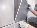 Person setzt gro&szlig;e graue Wandfliese mit Kleber ein, tr&auml;gt Handschuhe, pr&auml;zise Arbeit bei Fliesenverlegung sichtbar