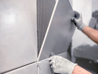 Person setzt gro&szlig;e graue Wandfliese mit Kleber ein, tr&auml;gt Handschuhe, pr&auml;zise Arbeit bei Fliesenverlegung sichtbar