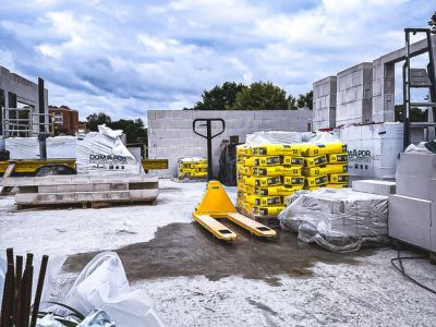 Baustelle mit Paletten voller Zements&auml;cke und Porenbetonbl&ouml;cken, Hubwagen steht bereit unter Wolkenhimmel.