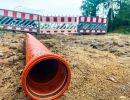 Orange Rohrleitung liegt auf sandigem Boden vor Baustellenabsperrung mit rot-wei&szlig;en Barrieren.