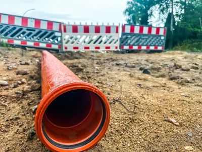 Orange Rohrleitung liegt auf sandigem Boden vor Baustellenabsperrung mit rot-wei&szlig;en Barrieren.