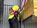 Person mit Warnweste und pinkem Auffanggurt arbeitet sicher neben Holzlager &ndash; zeigt Schutzma&szlig;nahmen im Industriebereich