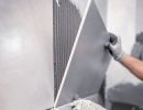 Person setzt gro&szlig;e graue Wandfliese mit Kleber ein, tr&auml;gt Handschuhe, pr&auml;zise Arbeit bei Fliesenverlegung sichtbar.